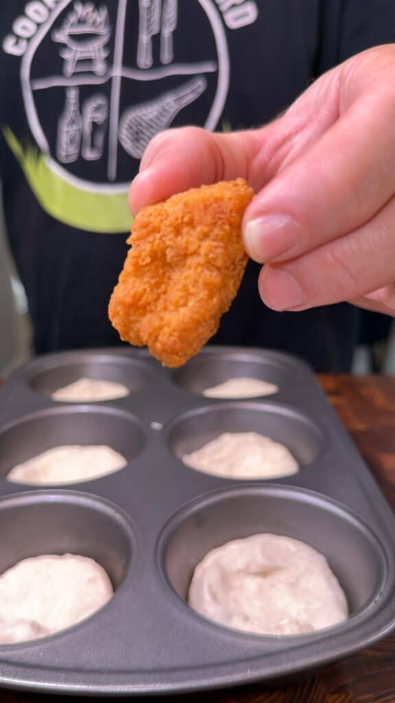 A hand holds a breaded chicken nugget over a muffin tin filled with biscuit dough.