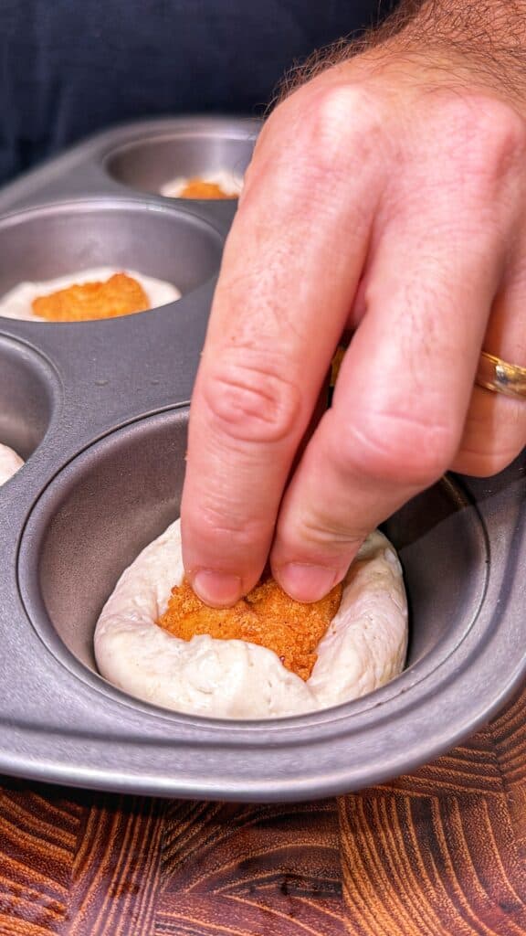 A hand presses a breaded chicken nugget into biscuit dough in a muffin tin.