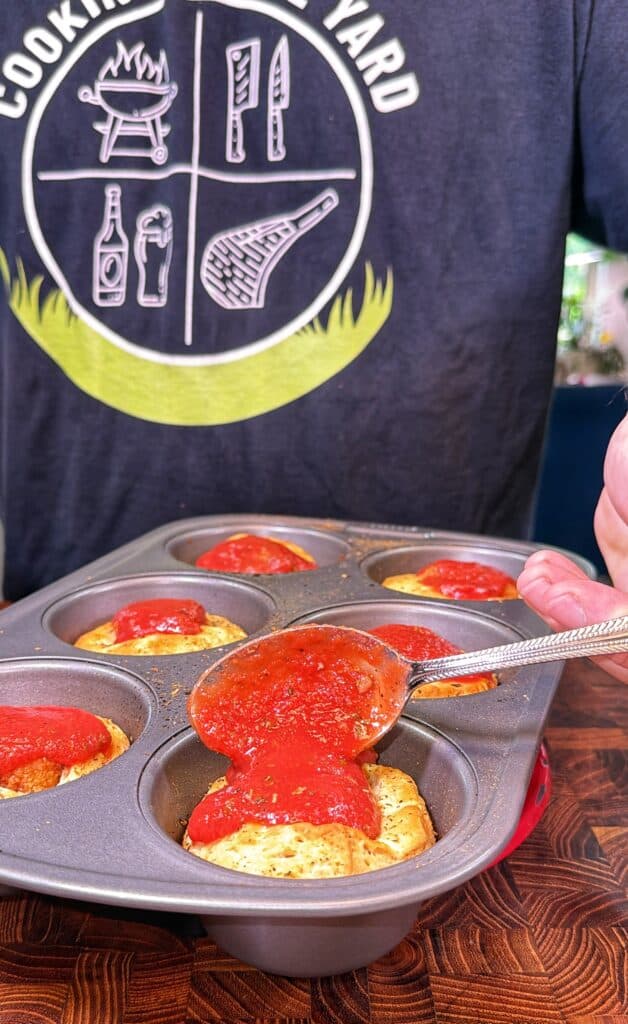 Red pizza sauce is spooned over baked biscuit cups in a muffin tin.