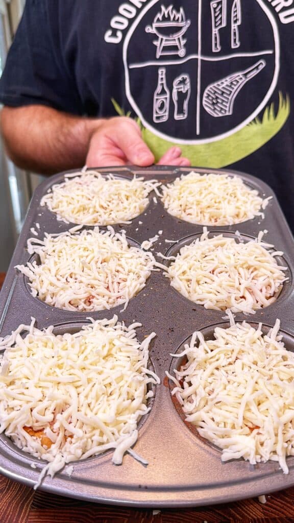A muffin tin holds six biscuit cups topped with shredded cheese, ready to go under the broiler.