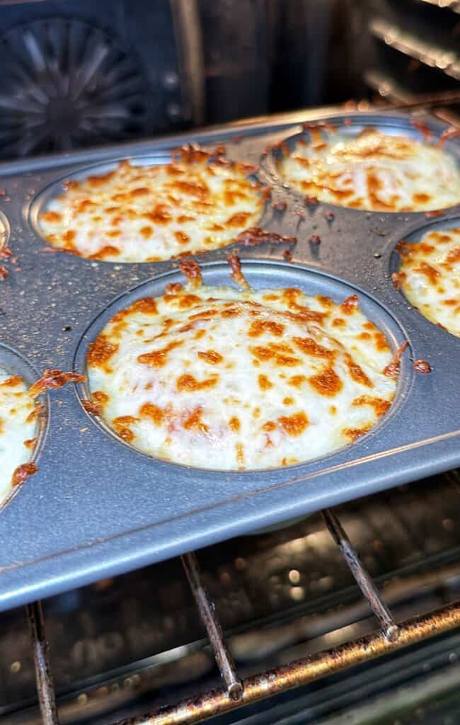 Chicken Parmesan biscuit bites topped with bubbly melted cheese bake in a muffin tin inside an oven.