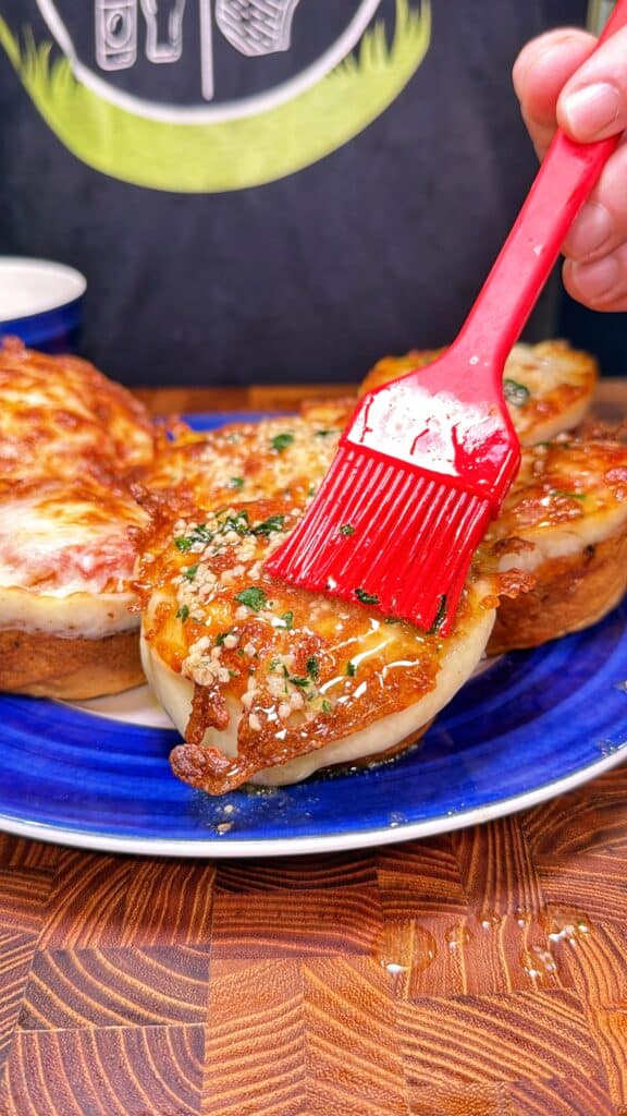 A hand uses a red brush to apply seasoning to chicken Parmesan biscuit bites on a blue plate.