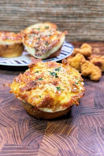 A cheesy chicken Parmesan biscuit bite topped with herbs sits on a wooden surface, with more biscuit bites and breaded chicken nuggets in the background.