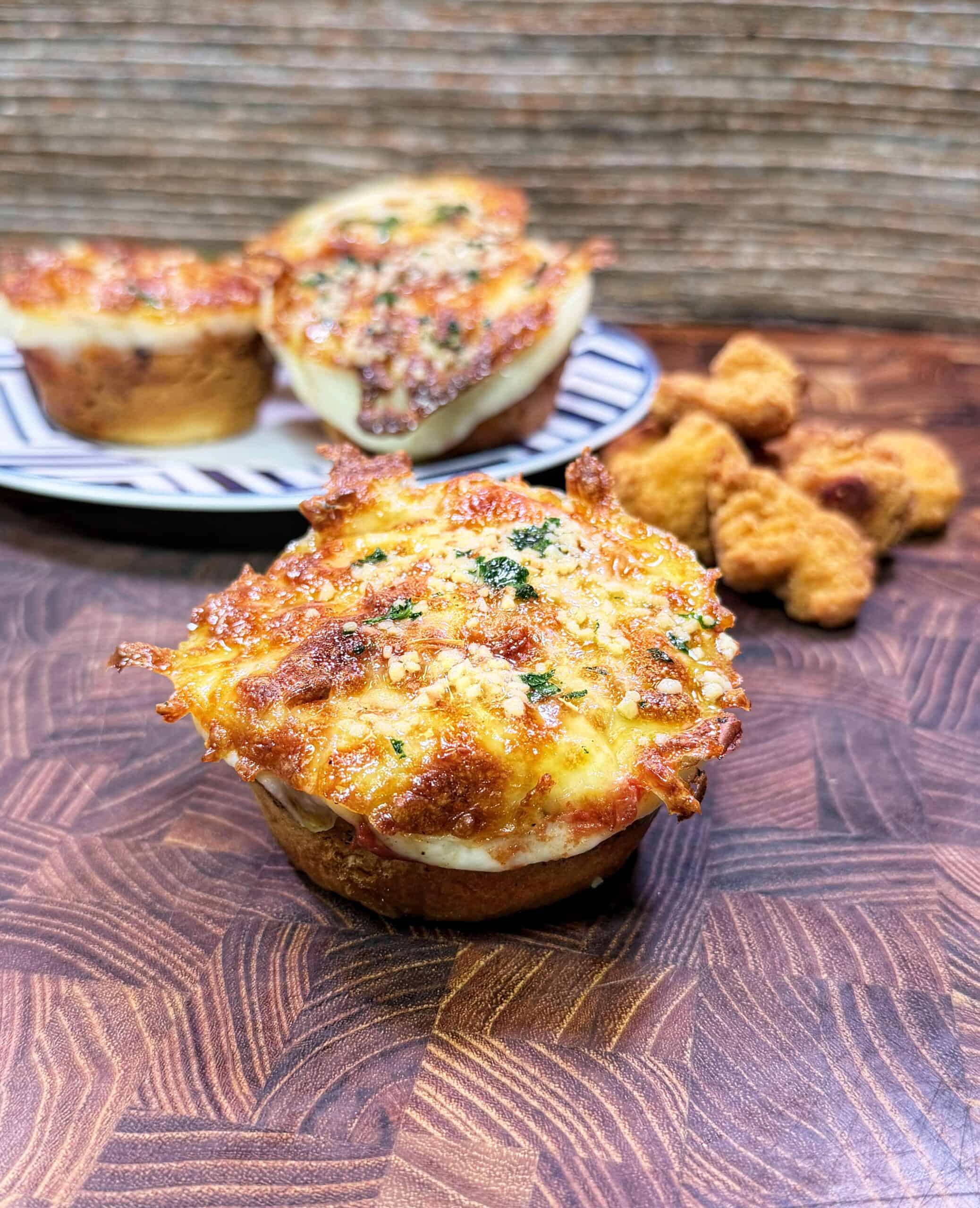 A cheesy chicken Parmesan biscuit bite topped with herbs sits on a wooden surface, with more biscuit bites and breaded chicken nuggets in the background.