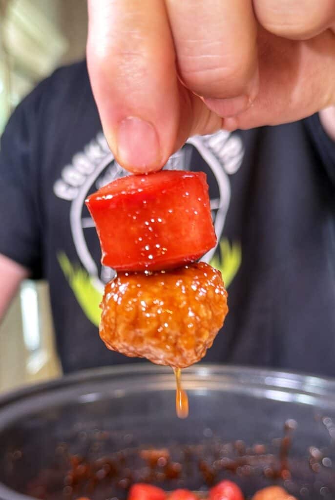 A hand holds a toothpick with a meatball and hot dog piece above a pot, with sauce dripping off.