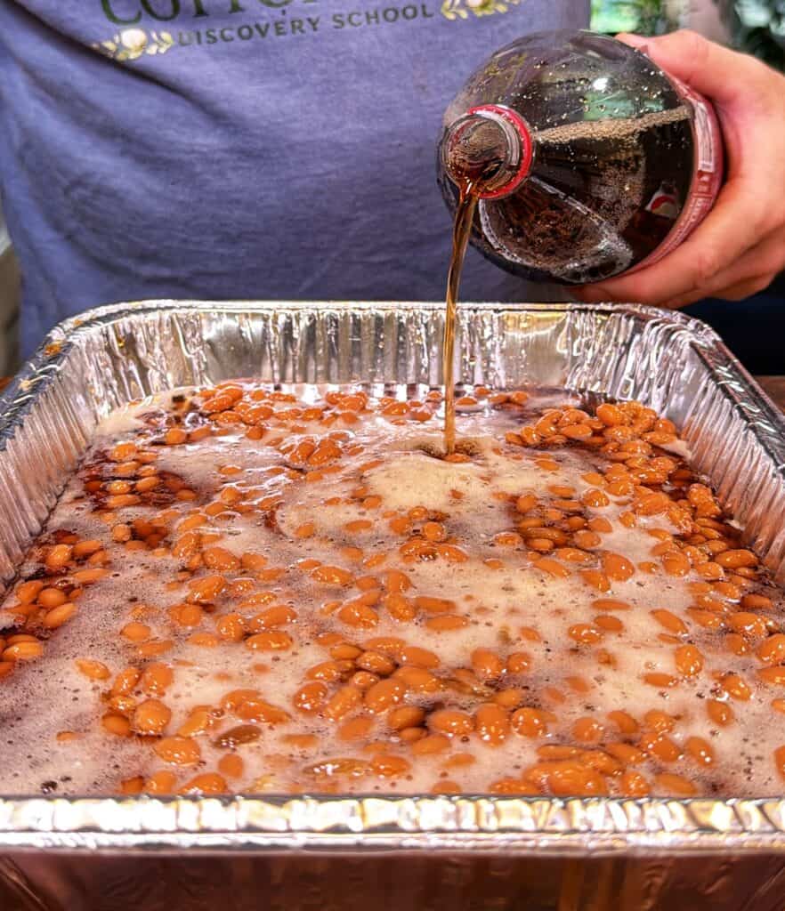 Dr Pepper is poured from a bottle into a foil tray of baked beans.