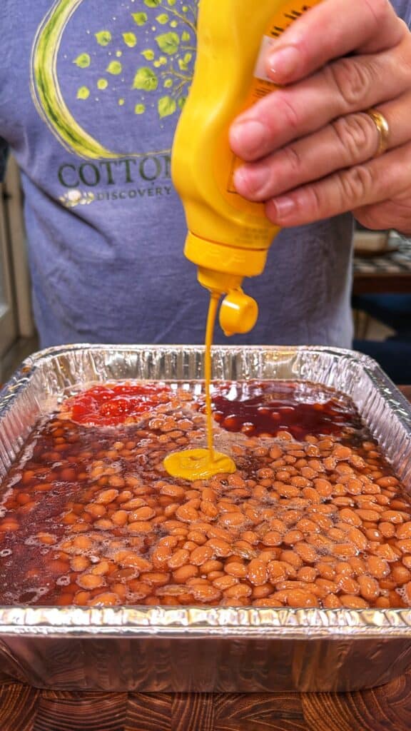 Yellow mustard is squeezed onto baked beans in an aluminum tray, with ketchup visible on top.