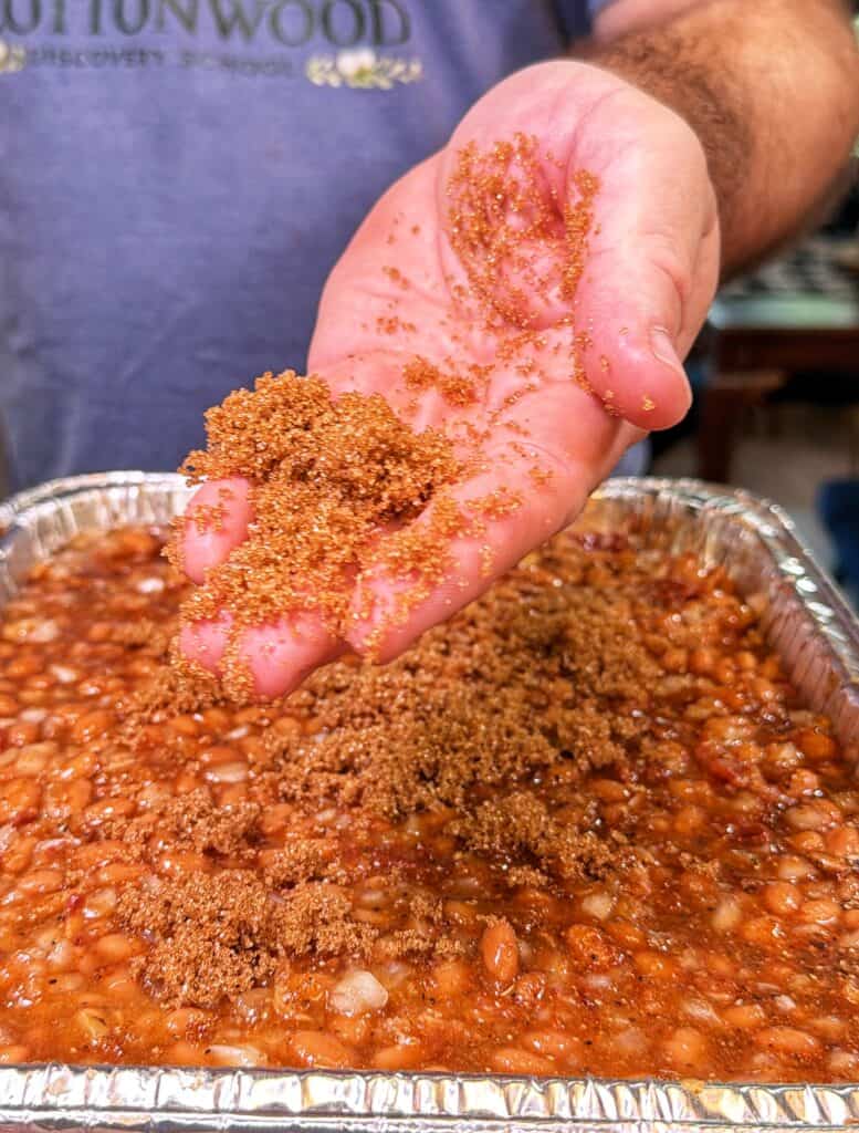 Brown sugar is sprinkled over baked beans in a foil tray.