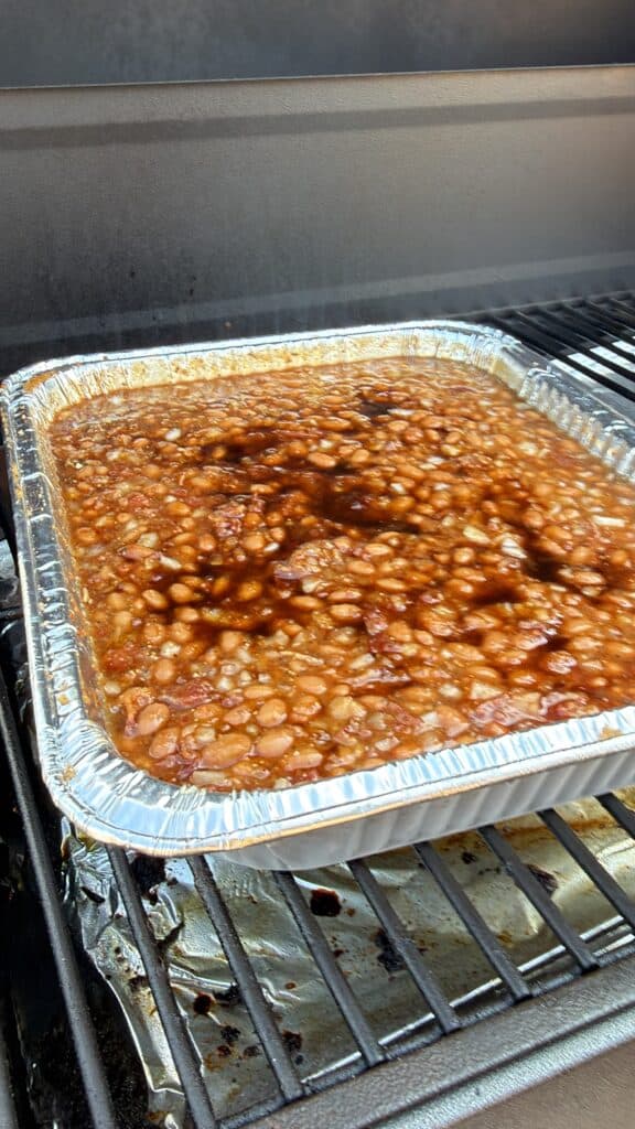 A foil tray of baked beans sits on a grill.