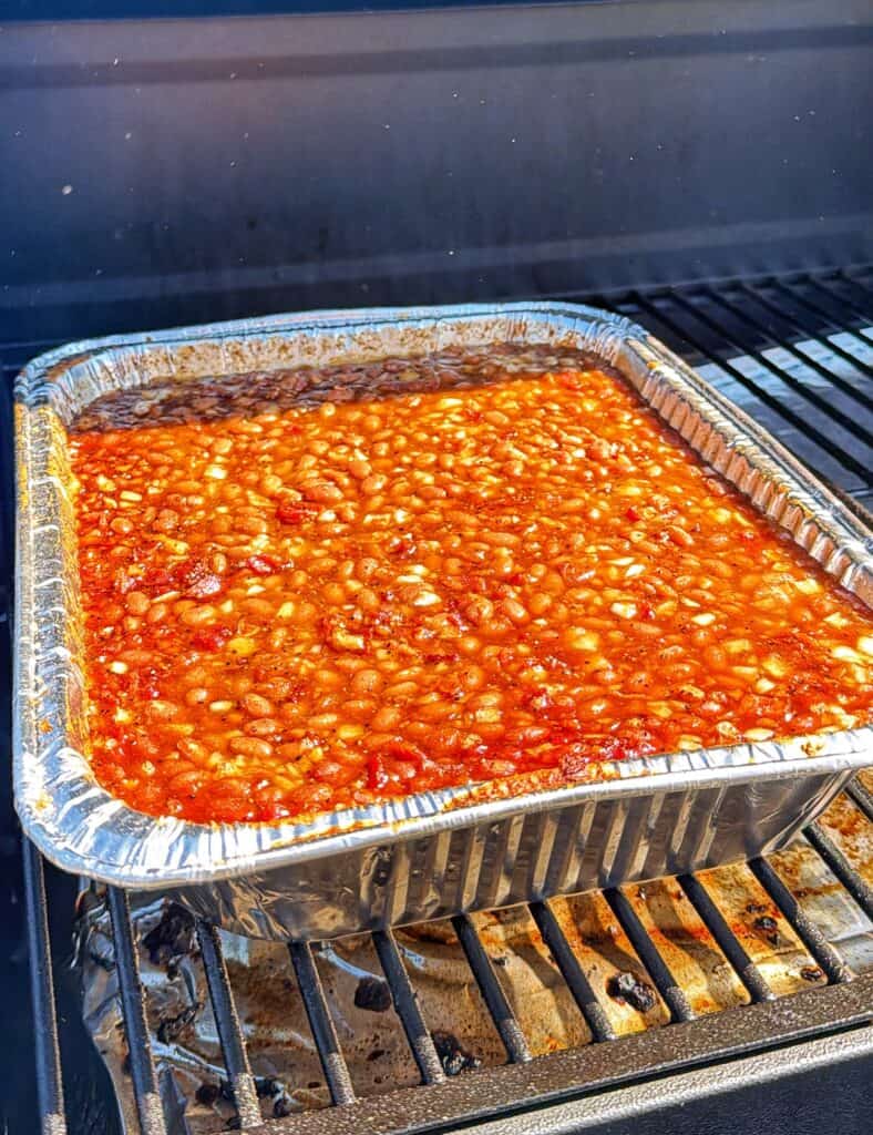 A foil tray of baked beans in sauce sits on a barbecue grill.