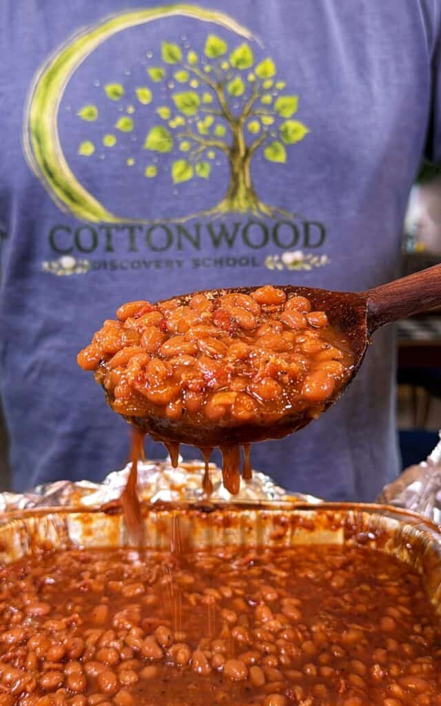 A spoonful of Dr Pepper baked beans is held over a pan, with sauce dripping back into the beans.