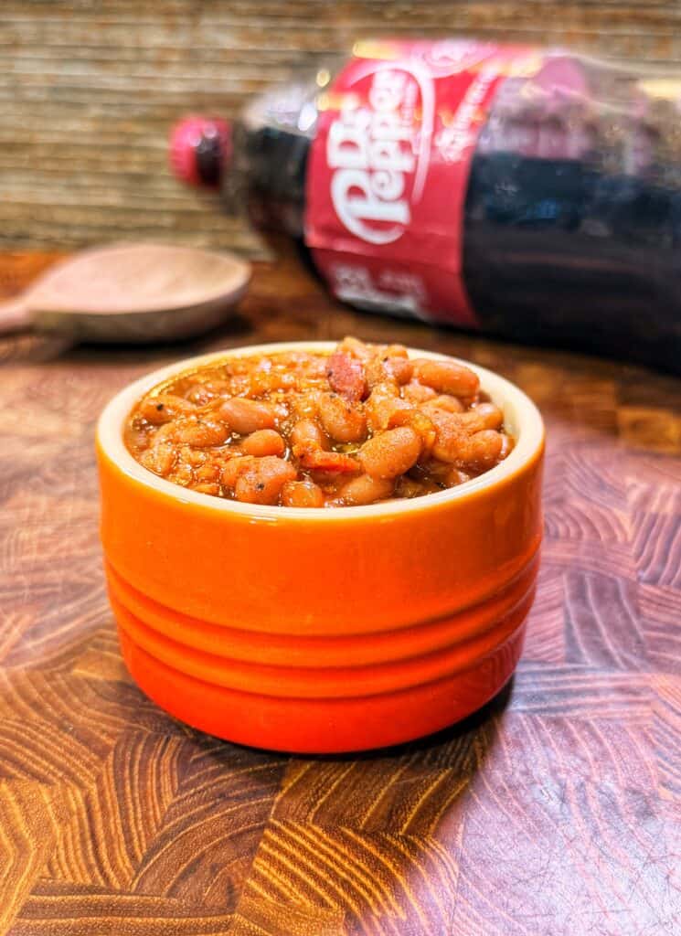 An orange bowl of Dr Pepper baked beans sits on a wooden table, with a wooden spoon and a Dr Pepper bottle in the background.