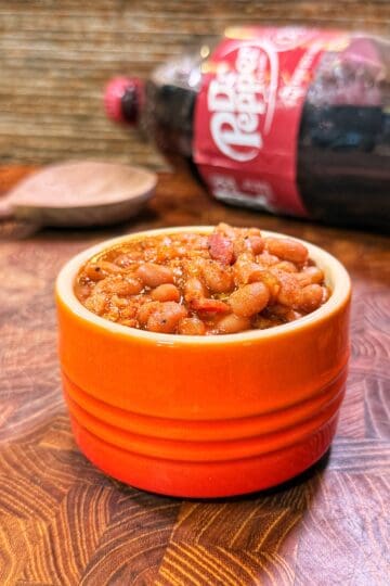 A bowl of Dr Pepper baked beans on a wooden surface, with a wooden spoon and a Dr Pepper bottle in the background.