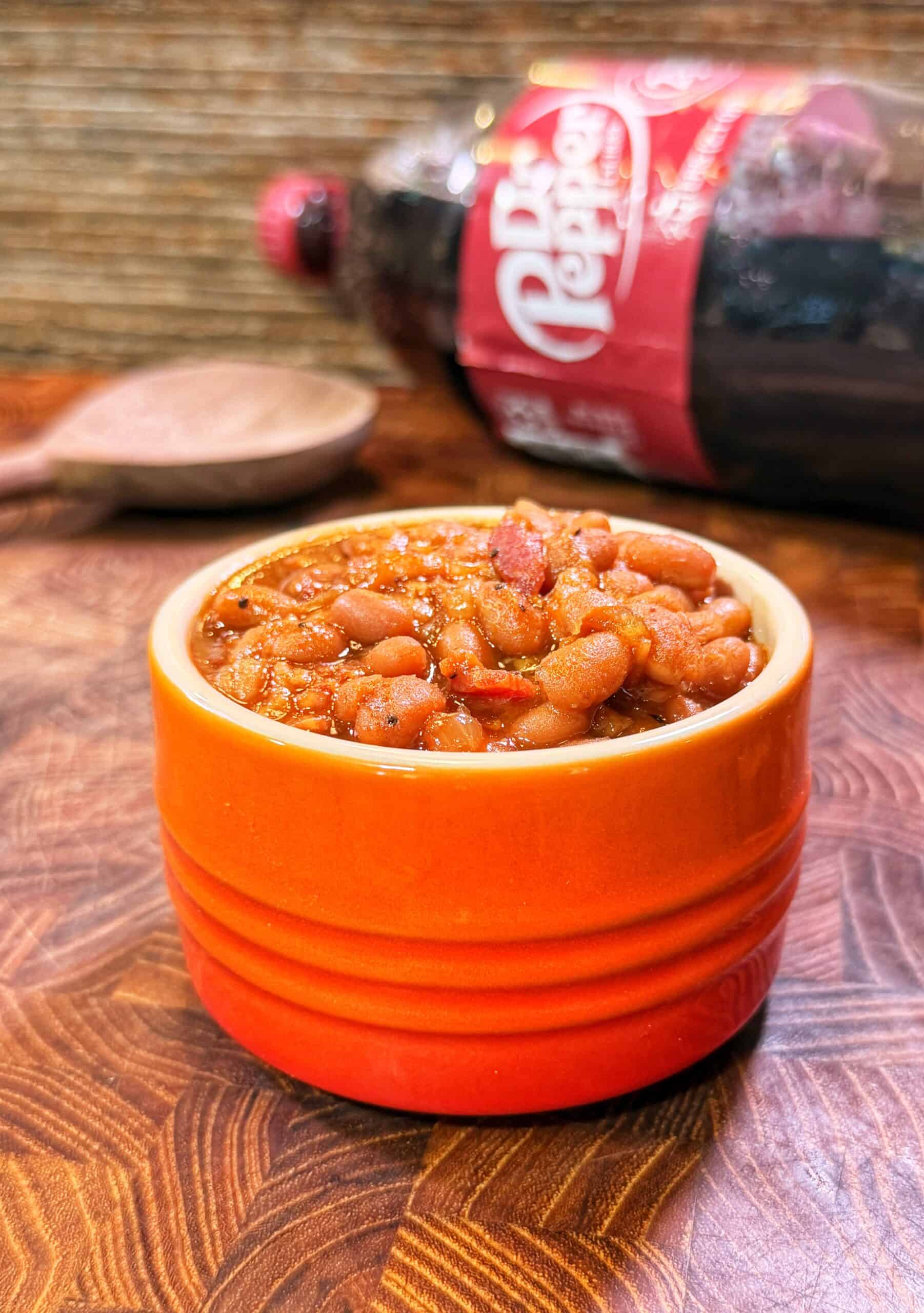 A bowl of Dr Pepper baked beans on a wooden surface, with a wooden spoon and a Dr Pepper bottle in the background.