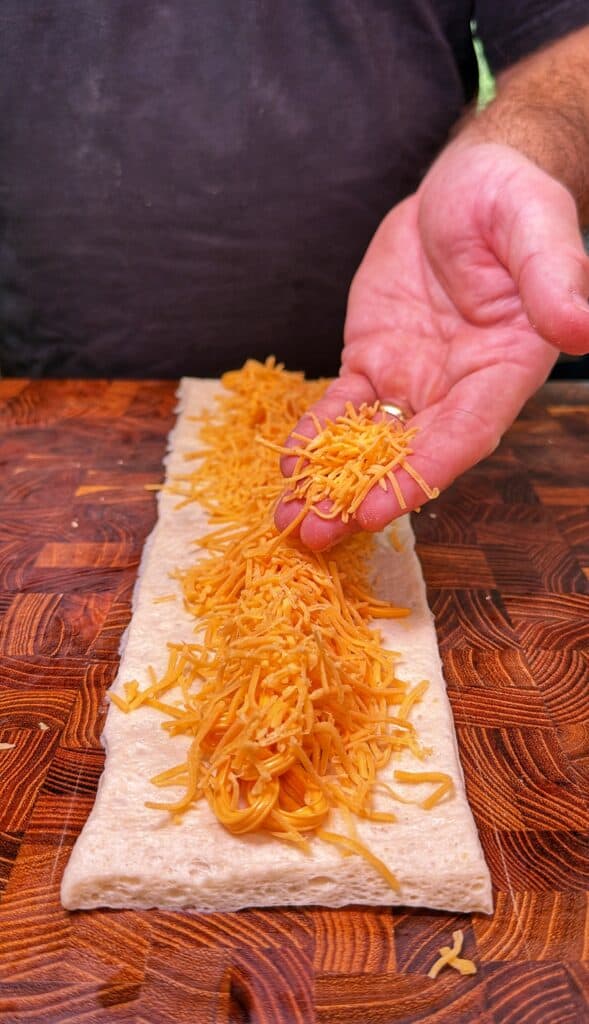 A hand sprinkles shredded cheddar cheese over a strip of dough.