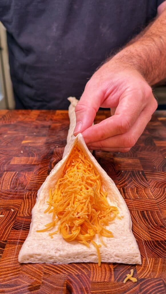 A person folds dough around shredded cheddar cheese to enclose the filling.