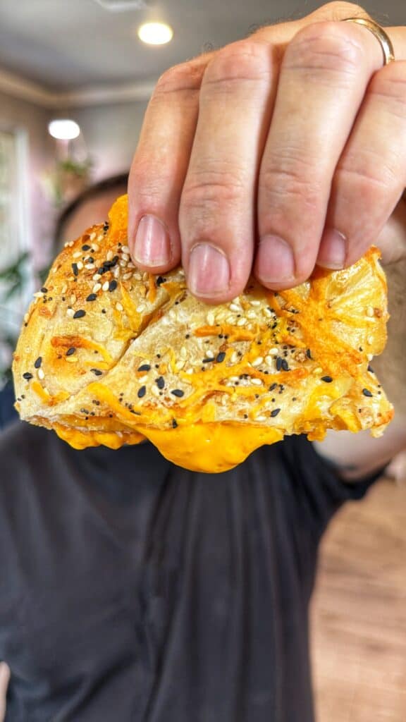 A hand holds a cheddar cheese stuffed pretzel with melted cheese in the center.