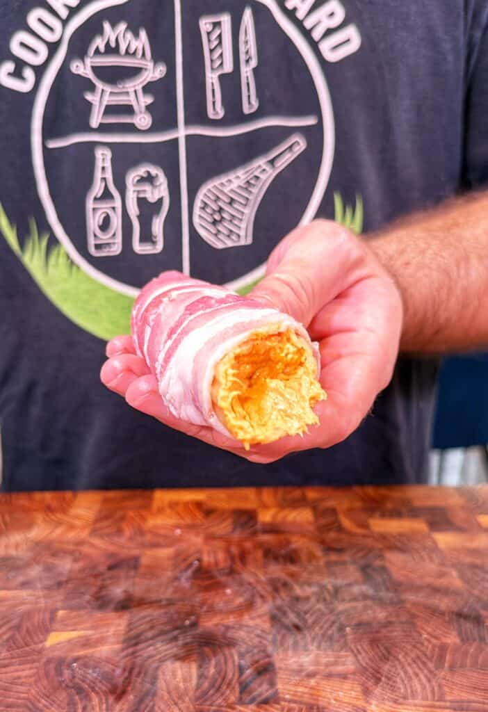 A hand holds a bacon-wrapped Buffalo Chicken Shotgun Shell with cheese filling visible.