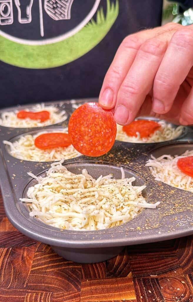 A hand places a pepperoni slice on shredded cheese in a muffin tin.