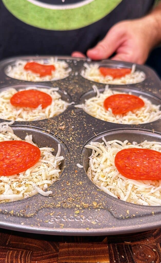 A person holds a muffin tin filled with shredded cheese and pepperoni-topped biscuit cups.