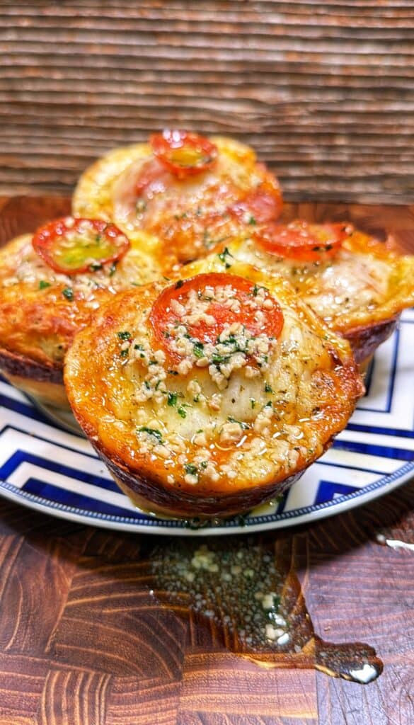 Four pepperoni pizza biscuit muffins topped with melted cheese and herbs on a patterned plate.