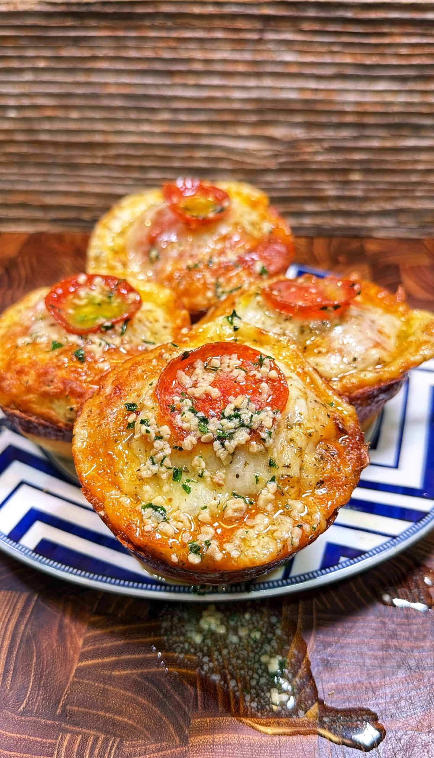 Four pepperoni pizza biscuit muffins topped with melted cheese, herbs, and garlic on a patterned plate.