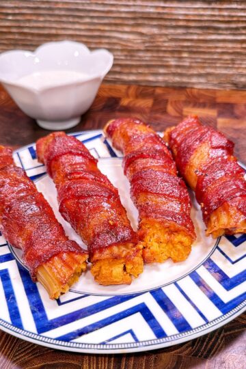 Four bacon-wrapped Buffalo Chicken Shotgun Shells on a blue and white plate with a small bowl of dipping sauce.