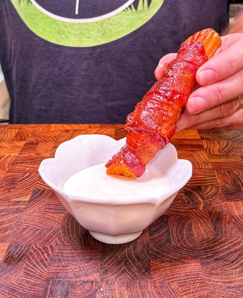 A hand dips a bacon-wrapped Buffalo Chicken Shotgun Shell into white dipping sauce.