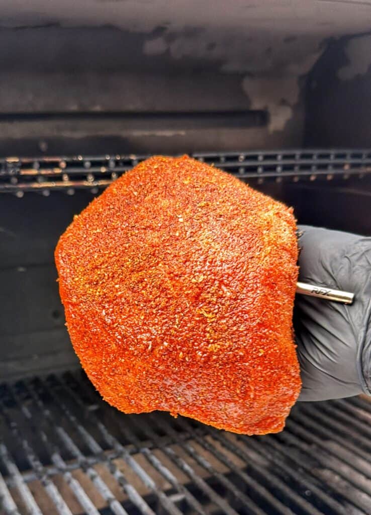 Gloved hand holding a seasoned turkey breast with a probe inserted near a smoker.