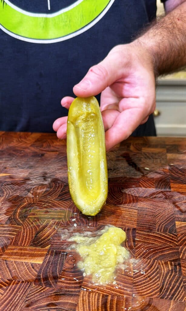 A hand holds a halved pickle with the center scooped out on a wooden cutting board.