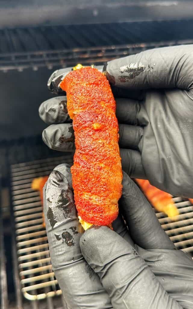 A hand holds a bacon-wrapped Buffalo Chicken Pickle Popper covered in red seasoning, with more poppers on a grill in the background.