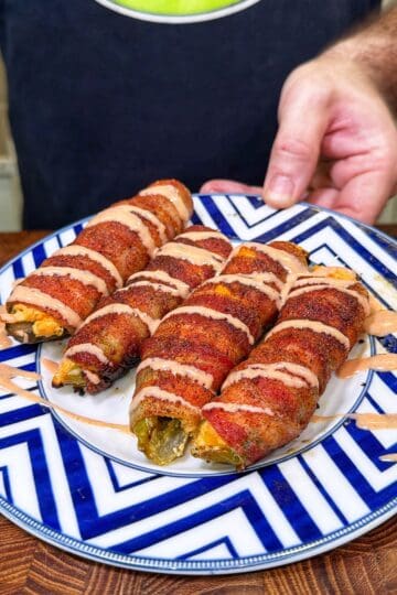 Four bacon-wrapped Buffalo Chicken Pickle Poppers drizzled with sauce on a blue and white zigzag-patterned plate.