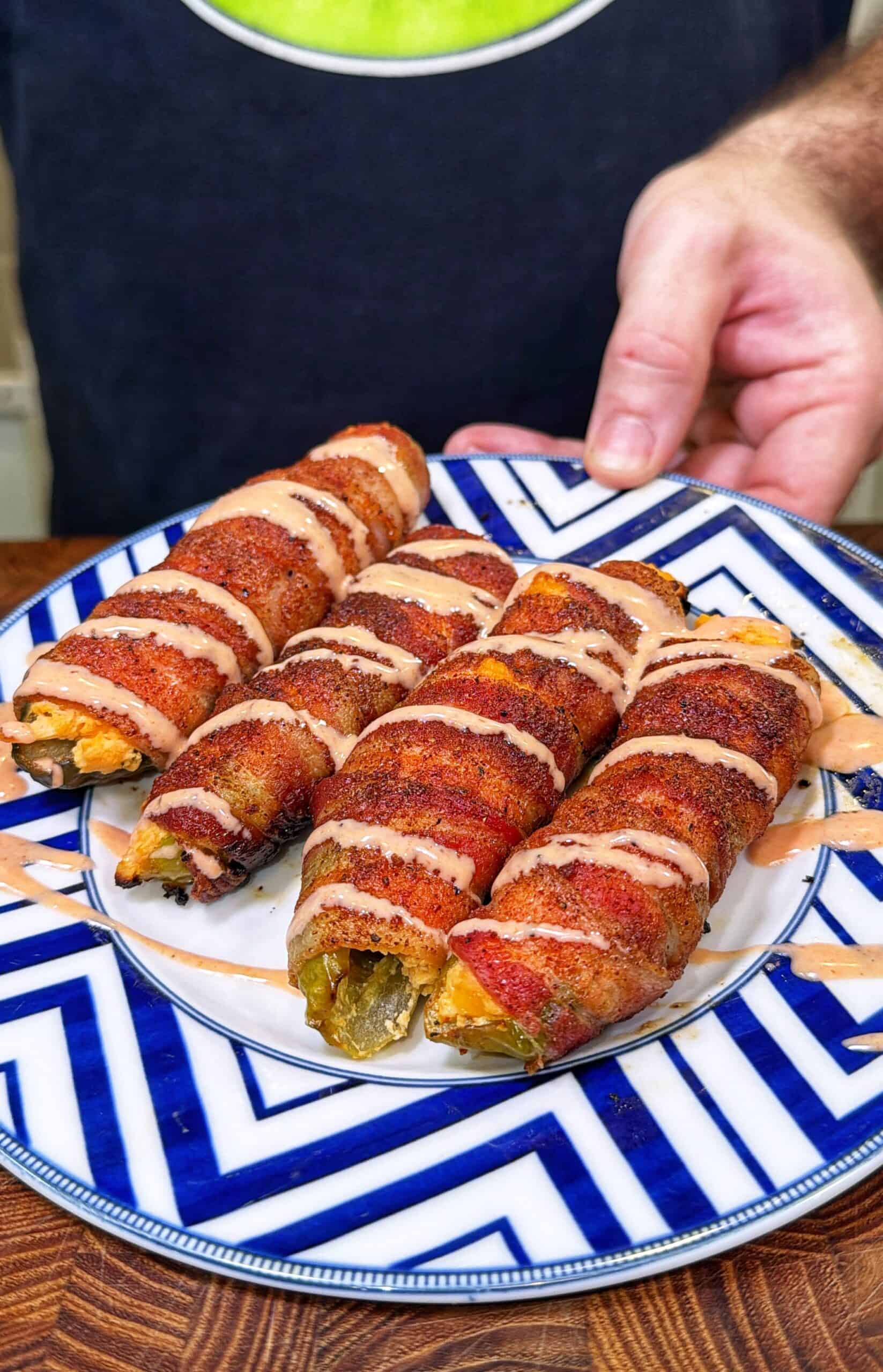 Four bacon-wrapped Buffalo Chicken Pickle Poppers drizzled with sauce on a blue and white zigzag-patterned plate.