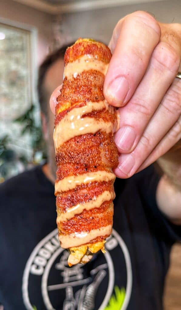 A Buffalo Chicken Pickle Popper covered in spices and drizzled with creamy sauce, held up indoors.