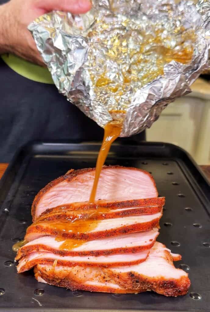 Sliced smoked turkey breast on a tray as a person pours glaze from foil over it.