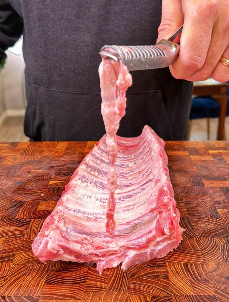 A person removes the membrane from a raw rack of ribs on a wooden cutting board using a metal tool.