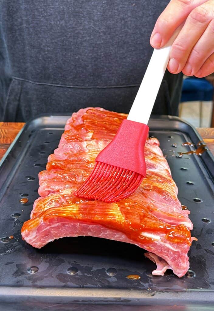 A person brushes sauce onto a raw rack of ribs on a black tray using a red silicone brush.