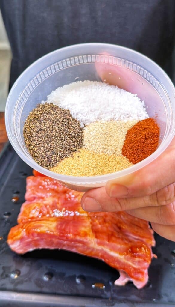 A hand holds a container of seasonings above raw ribs on a black tray.