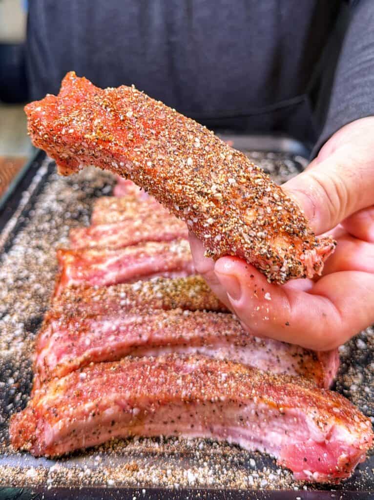 A hand holds a seasoned raw rib above a tray of ribs coated in a peppery rub.