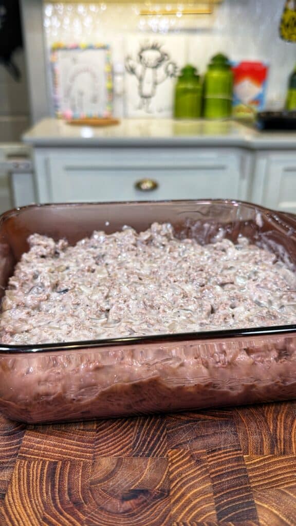 A square glass baking dish filled with a creamy cooked ground beef mixture on a wooden cutting board.