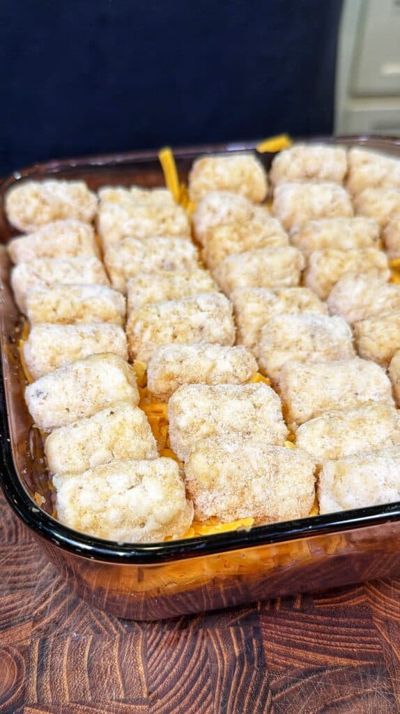 A glass casserole dish with shredded cheddar cheese topped with frozen tater tots.