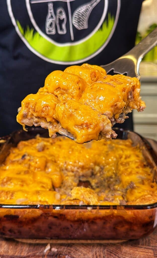 A metal spatula lifting a serving of cheeseburger tater tot casserole from a glass baking dish.