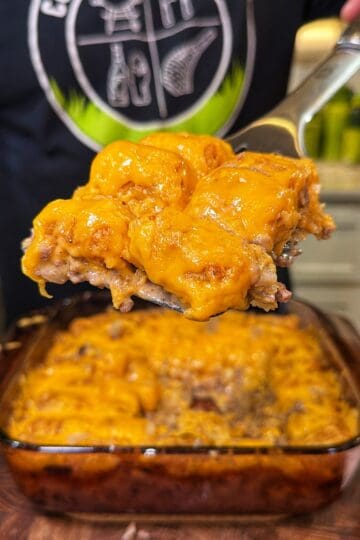 A metal spatula lifting a serving of cheeseburger tater tot casserole from a glass baking dish.
