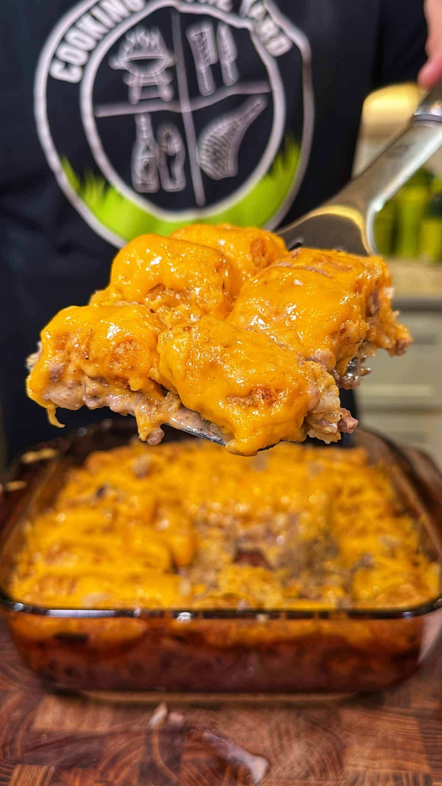 A metal spatula lifting a serving of cheeseburger tater tot casserole from a glass baking dish.