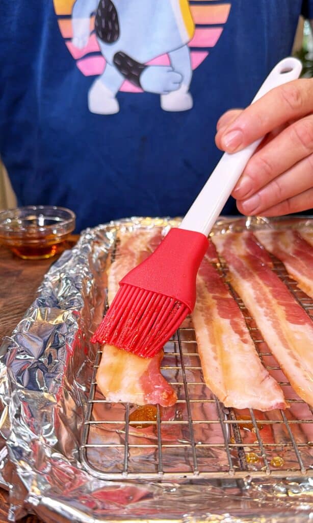 A hand brushes maple syrup over raw bacon strips on a foil-lined rack.