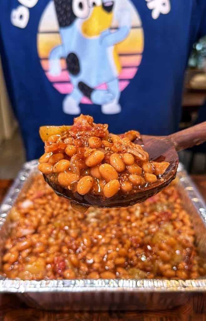 Wooden spoon holding apple pie baked beans over a tray of beans.