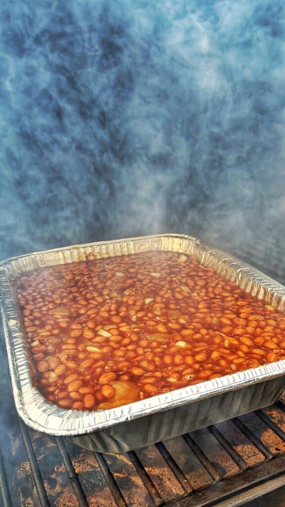 Foil tray of apple pie baked beans smoking on a grill.