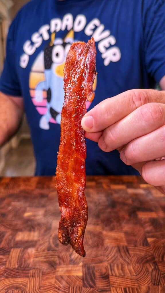 A hand holds a crispy bacon strip over a wooden surface.
