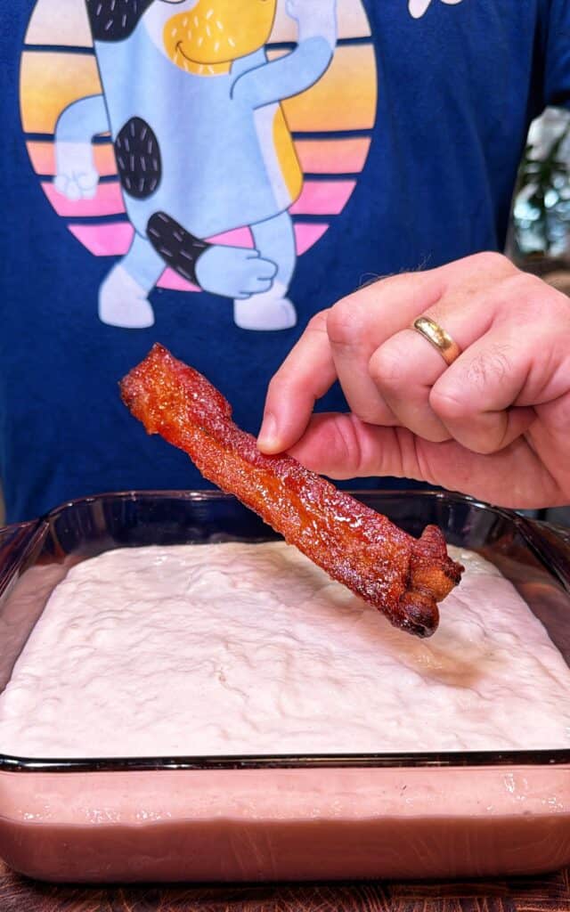 A hand holds a bacon strip above a glass dish of pancake batter.