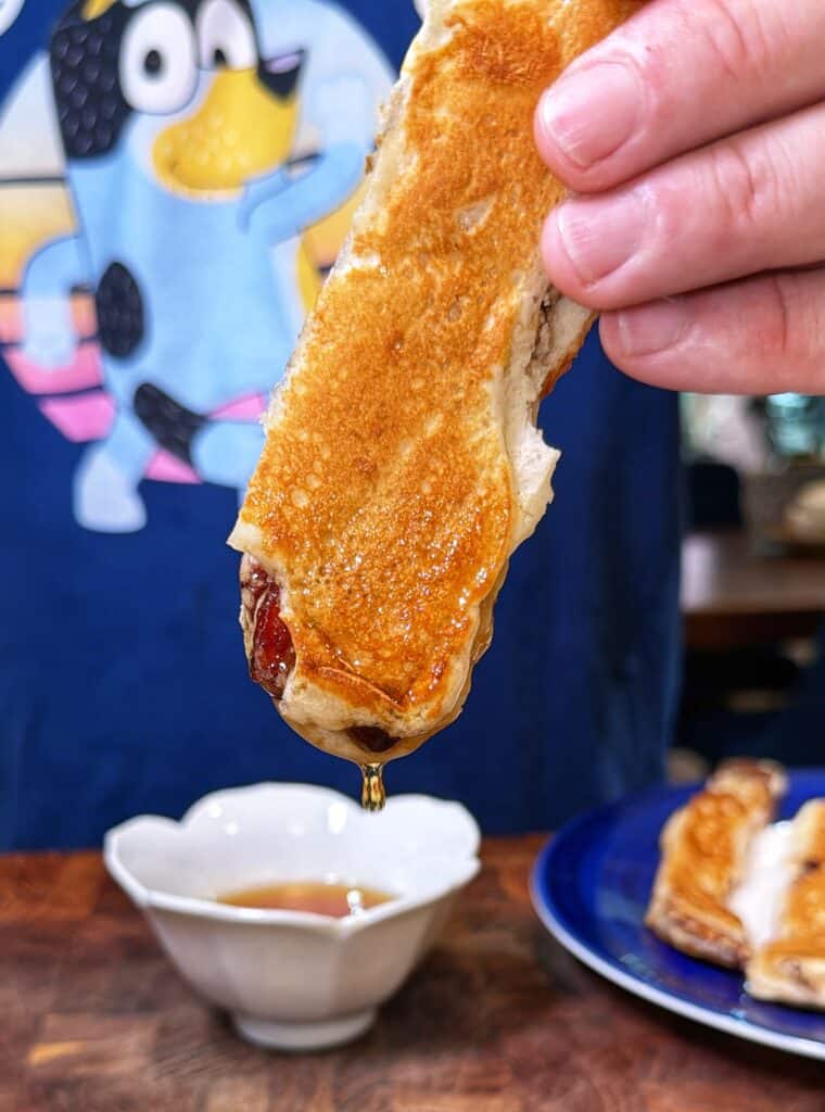 A hand dips a pancake-battered bacon strip into syrup.